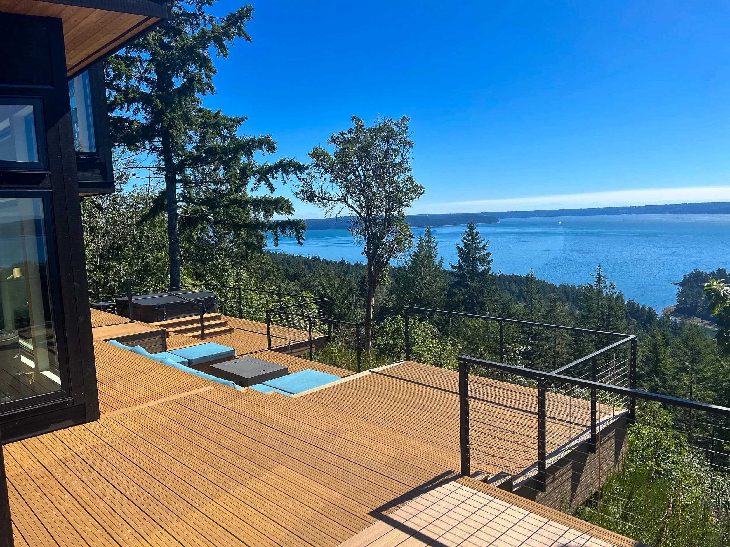 A multi-level Sylvanix composite deck with cable railings and a hot tub, overlooking forest and open water.